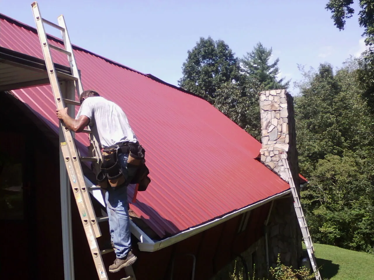 Roofer on a ladder beside a red metal roof and stone chimney — Soffit & Fascia Repair by Roofing Brothers in Balko